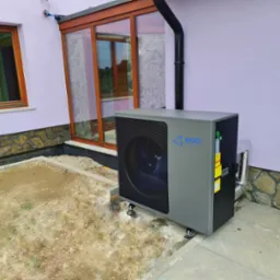 Szara pompa ciepła ED030-HCD z niebieskim logo, zamontowana na zewnątrz budynku z fioletową elewacją i kamiennym cokołem, widoczny fragment okna i przeszklonego ganku.