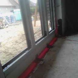 Montaż okna balkonowego z widocznym zabezpieczeniem w postaci czerwonej folii w płynie i metalowymi mocowaniami. Na zewnątrz widoczne materiały budowlane i przenośna toaleta.