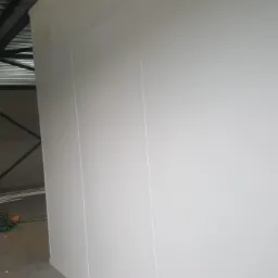 Wnętrze z wykończoną ścianą z białych paneli, widoczne łączenia między płytami i fragment konstrukcji dachu.