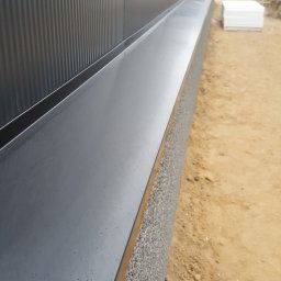 Mateusz Cybulski - Detal zdjęcia: Metalowa elewacja budynku z widocznym cokolem wykończonym szarą strukturą, sąsiadująca z piaszczystym terenem budowy.