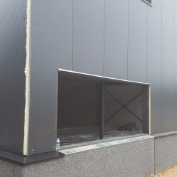 Mateusz Cybulski - Szary budynek przemysłowy w trakcie budowy z widocznymi otworami okiennymi i fundamentem wykończonym szarym kamieniem, butelka wody na parapecie