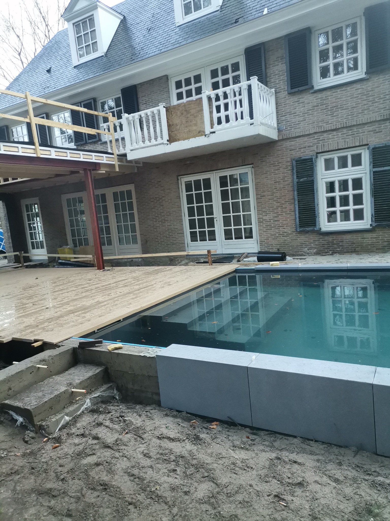 Budowa tarasu i basenu przy domu z cegły. Widoczne elementy konstrukcyjne, deski podłogowe i lustrzana tafla wody basenu.