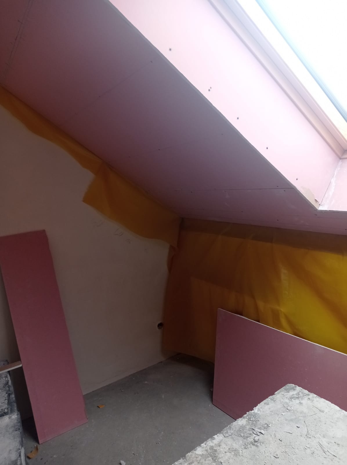 Montaż różowych płyt kartonowo-gipsowych na poddaszu ze skośnym sufitem, widoczna żółta folia paroizolacyjna oraz okno dachowe w trakcie prac wykończeniowych.