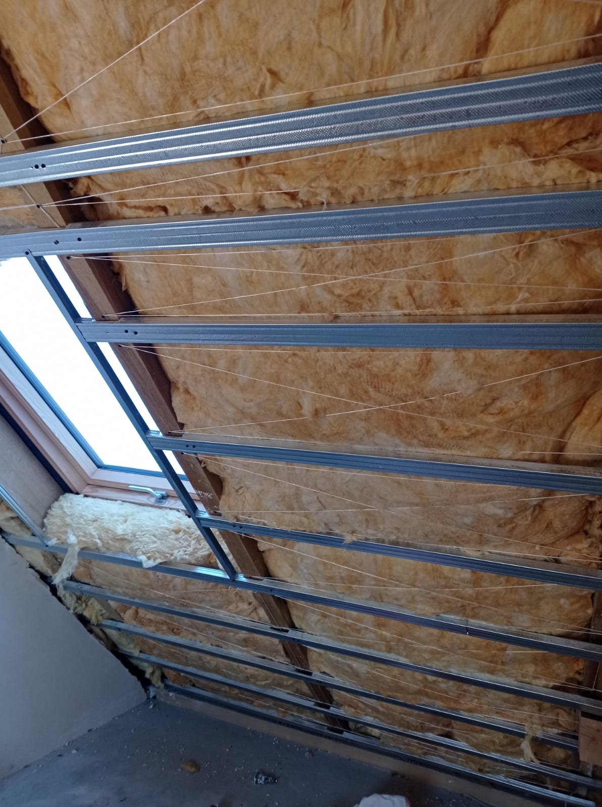 Skośny sufit poddasza z oknem dachowym, ocieplony wełną mineralną, z zamontowaną metalową konstrukcją pod płyty gipsowo-kartonowe, widok z dołu.