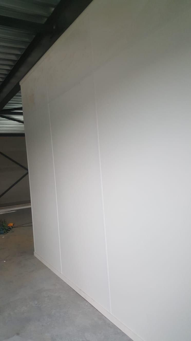Wnętrze z wykończoną ścianą z białych paneli, widoczne łączenia między płytami i fragment konstrukcji dachu.