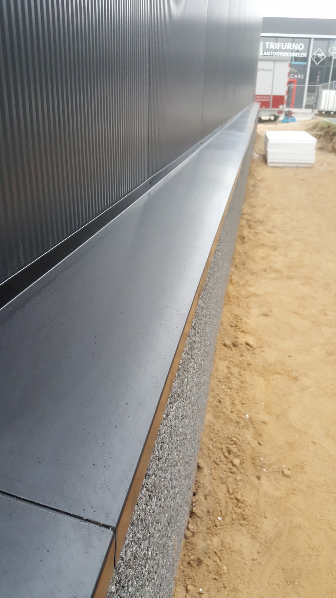 Detal zdjęcia: Metalowa elewacja budynku z widocznym cokolem wykończonym szarą strukturą, sąsiadująca z piaszczystym terenem budowy.