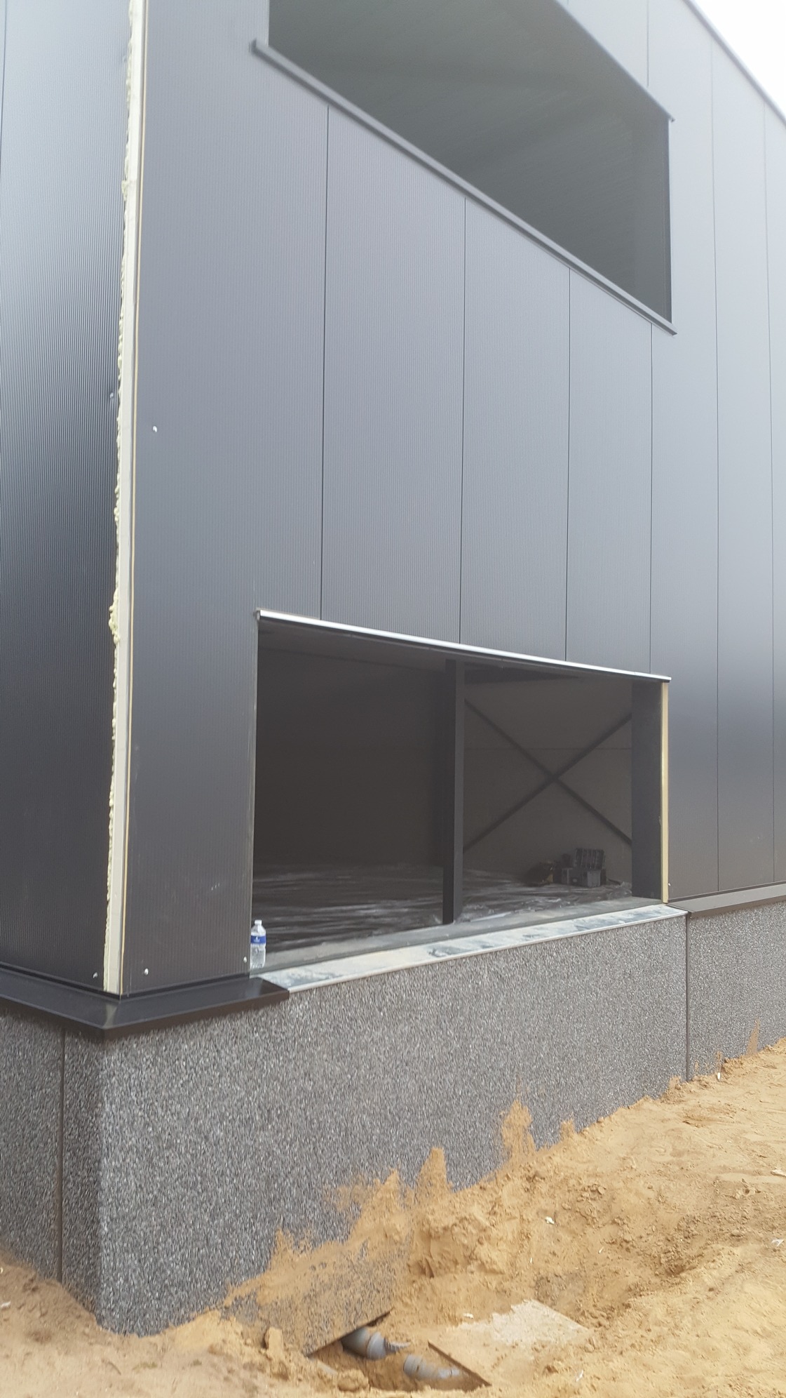 Szary budynek przemysłowy w trakcie budowy z widocznymi otworami okiennymi i fundamentem wykończonym szarym kamieniem, butelka wody na parapecie