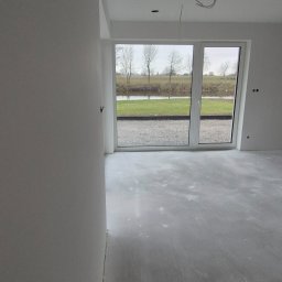 Best House - Surowe wnętrze z nałożonymi gładziami na ścianach, widoczne kable wystające z sufitu i duże okno z widokiem na zewnątrz.