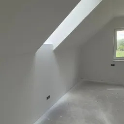 Poddasze po nałożeniu gładzi w Janowie Lubelskim. Widoczne okno dachowe, okno na ścianie, puszki elektryczne i stos paczek z materiałem wykończeniowym.