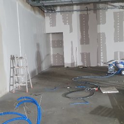 Best House - Wnętrze w trakcie prac wykończeniowych, ściany częściowo pokryte gładzią, widoczna maszyna do nakładania gładzi, drabina malarska i elementy instalacji elektrycznej na podłodze, pracownik w tle.