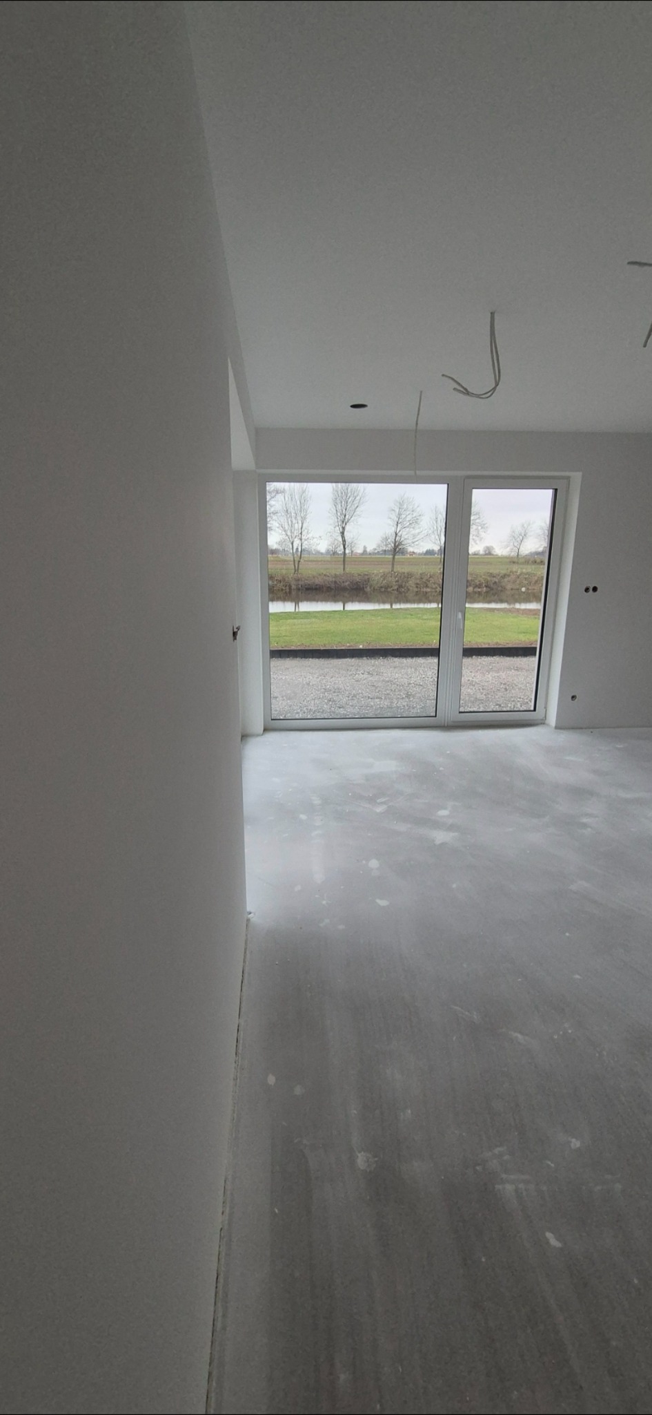 Surowe wnętrze z nałożonymi gładziami na ścianach, widoczne kable wystające z sufitu i duże okno z widokiem na zewnątrz.