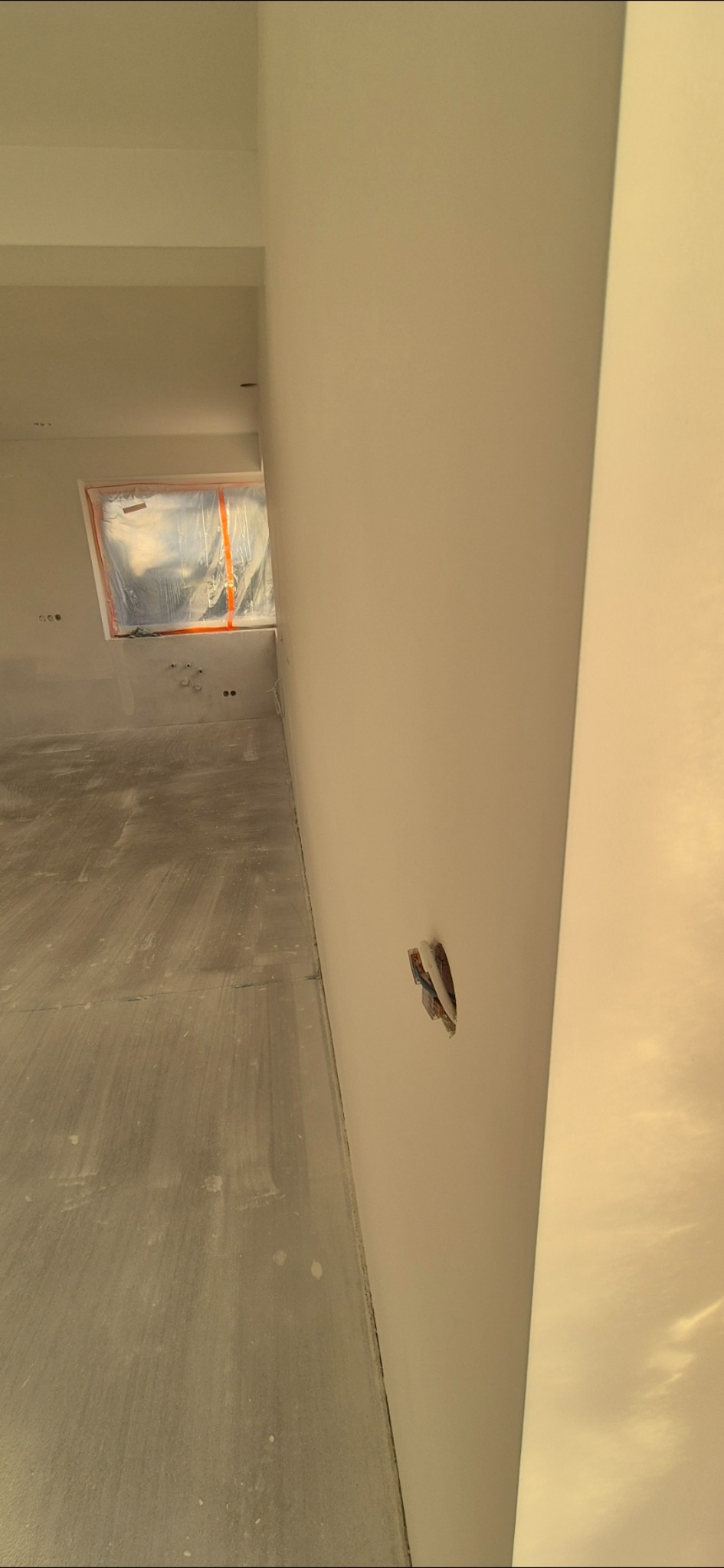 Świeżo wygładzona ściana z wystającą puszką elektryczną w pomieszczeniu w trakcie remontu, okno zabezpieczone folią malarską.