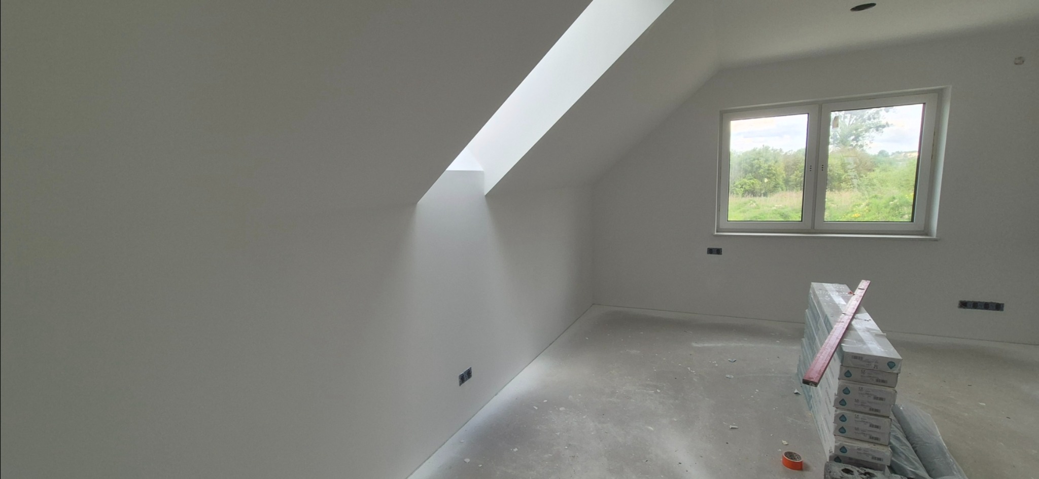 Poddasze po nałożeniu gładzi w Janowie Lubelskim. Widoczne okno dachowe, okno na ścianie, puszki elektryczne i stos paczek z materiałem wykończeniowym.