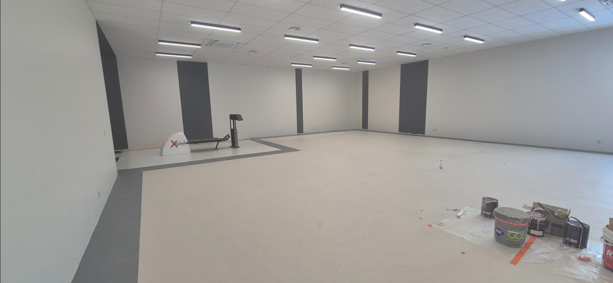 Jasne wnętrze sali z szarymi pasami na ścianach i sprzętem do ćwiczeń. Na podłodze widoczne puszki farb, co sugeruje prace wykończeniowe w toku.