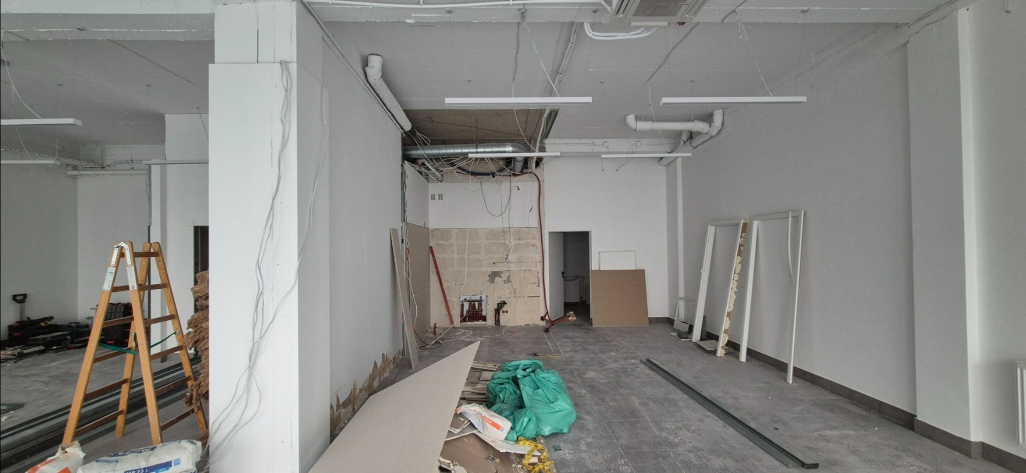 Wnętrze w trakcie remontu: ściany przygotowane do gładzi, widoczne instalacje, drabina, worki z materiałami budowlanymi. Surowy stan wykończenia.