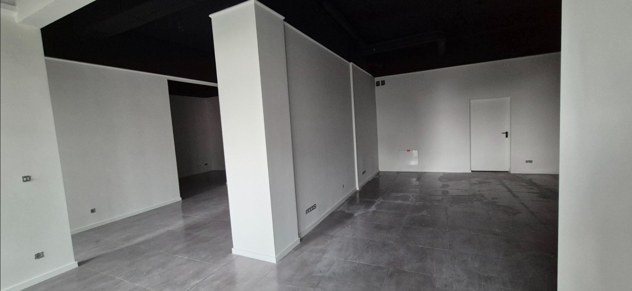 Wnętrze po remoncie: białe ściany, szara podłoga, czarny sufit. Widoczne gniazdka elektryczne i drzwi z czarną klamką. Minimalistyczny styl, świeżo po pracach wykończeniowych.