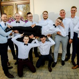 Kreatywna Fotografia Magdalena Saratowicz-BIałobok - Pan młody niesiony przez drużbów w białych koszulach i muszkach na tle białej ściany z oknem i kwiatami.