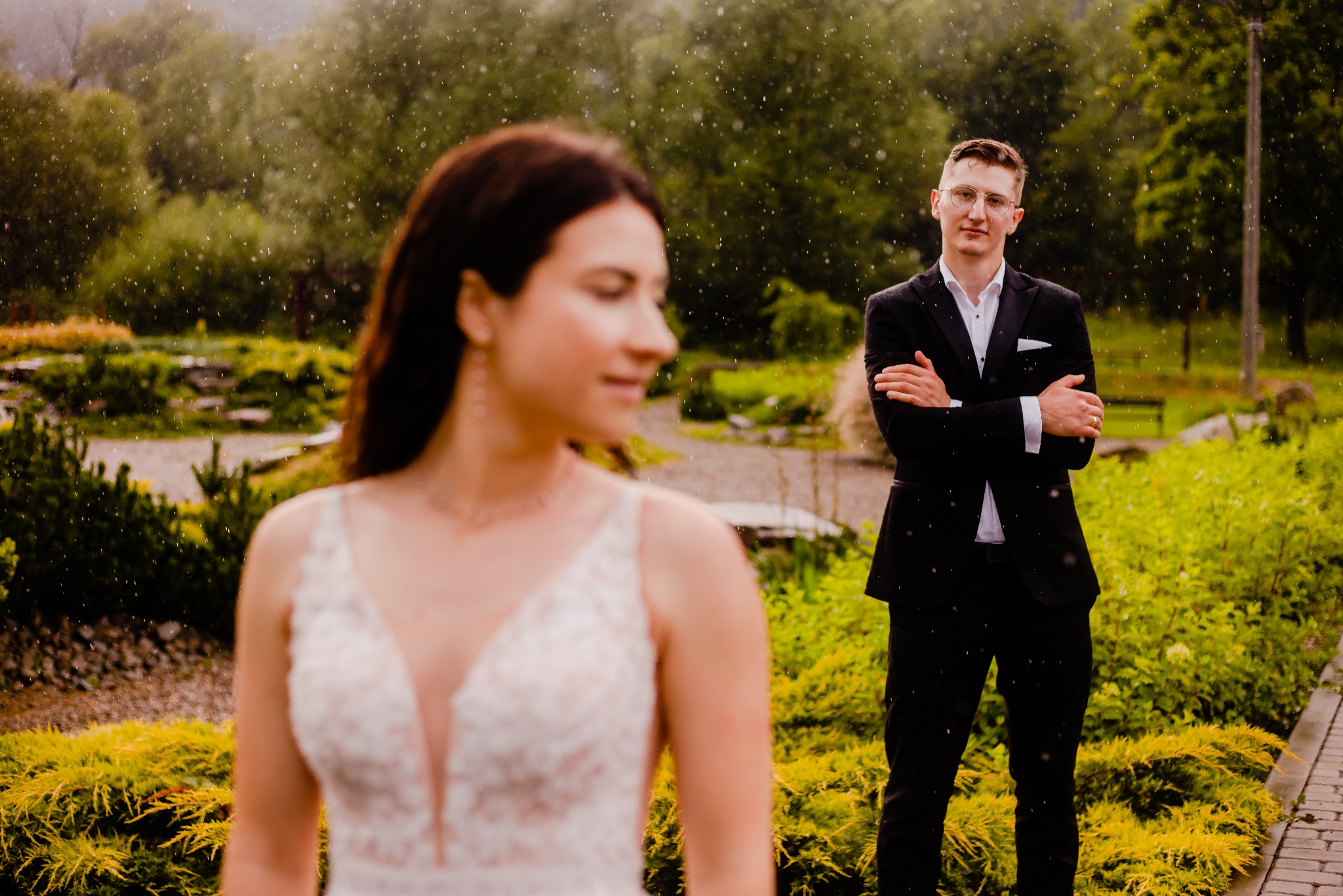 Para młoda w lekkim deszczu: panna młoda w koronkowej sukni z głębokim dekoltem patrzy w bok, pan młody w czarnym garniturze z założonymi rękami stoi w tle.
