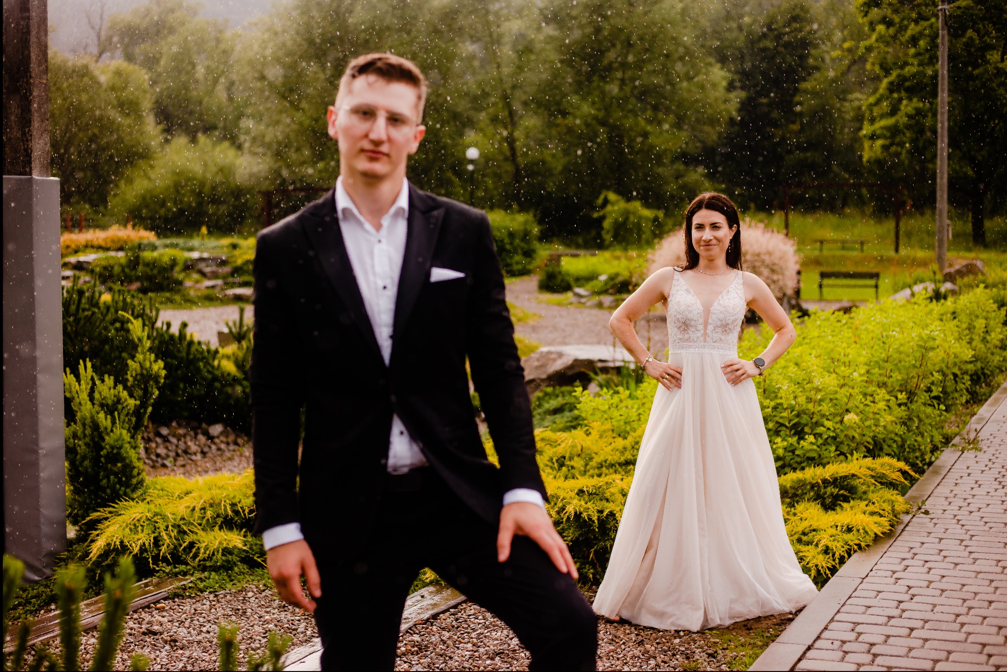 Para młoda w plenerze podczas deszczu: pan młody w czarnym garniturze z przodu, panna młoda w białej sukni z koronkową górą w tle, oboje uśmiechnięci.