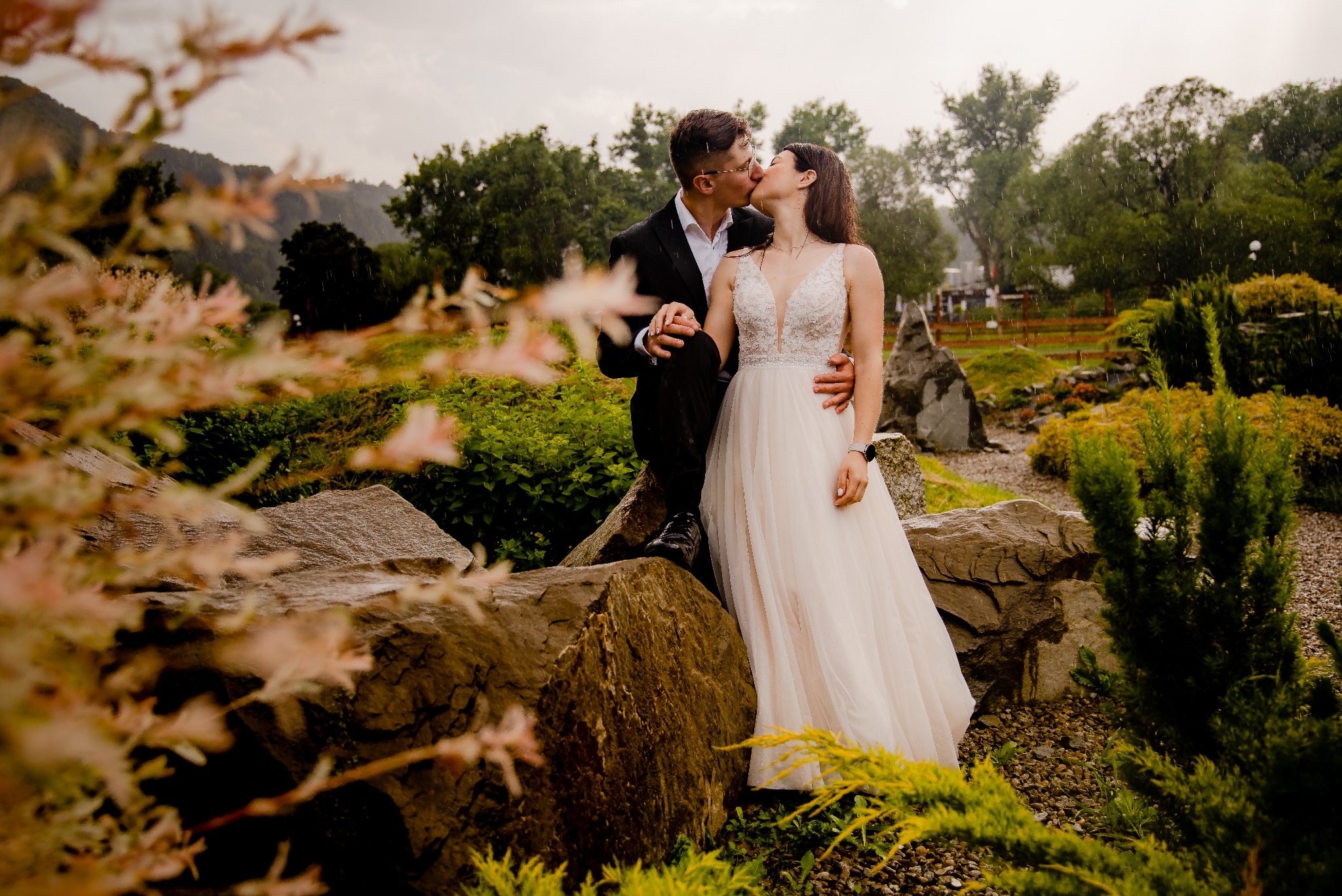 Para młoda całuje się na tle zieleni i skał w deszczowy dzień, panna młoda w białej sukni z koronkową górą, pan młody w czarnym garniturze i okularach.