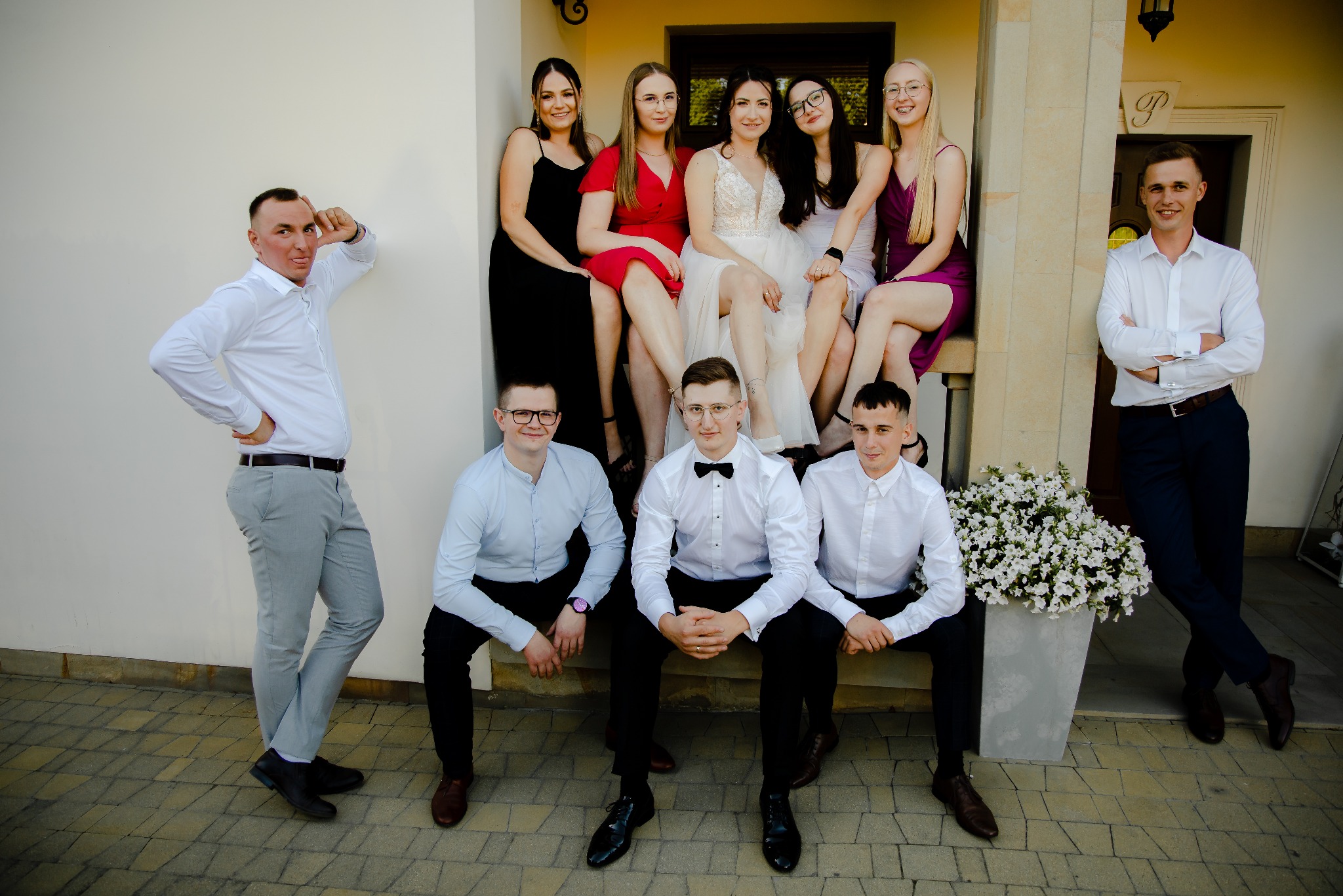 Grupa młodych ludzi, panna młoda w sukni ślubnej pośrodku, druhny w sukienkach i drużbowie w koszulach, pozują do zdjęcia przed budynkiem z kolumnami i kwiatami.