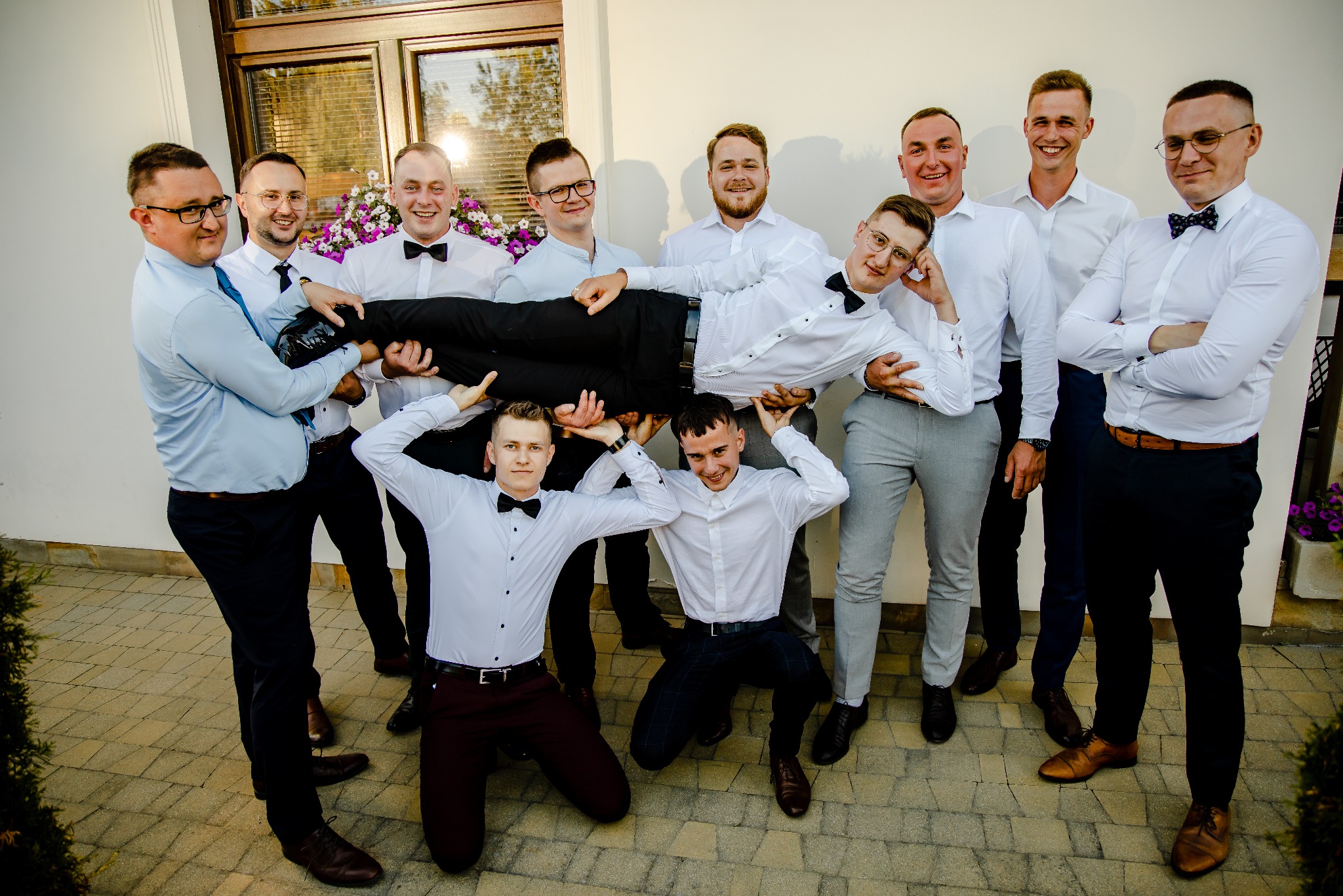 Pan młody niesiony przez drużbów w białych koszulach i muszkach na tle białej ściany z oknem i kwiatami.