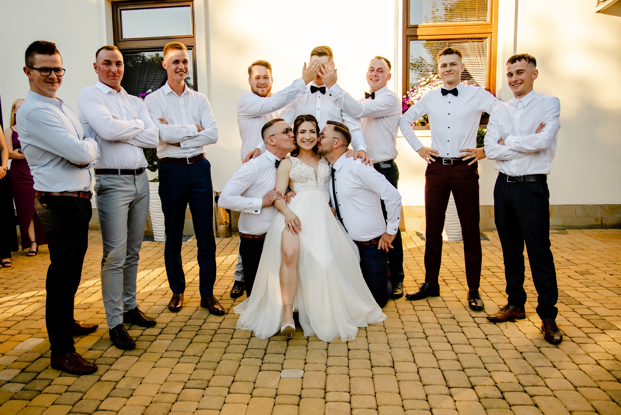 Panna młoda w białej sukni z rozcięciem na nodze, otoczona przez drużbów w białych koszulach i muchach, otrzymuje pocałunki w policzki od dwóch mężczyzn, podczas gdy jeden zasłania oczy,...