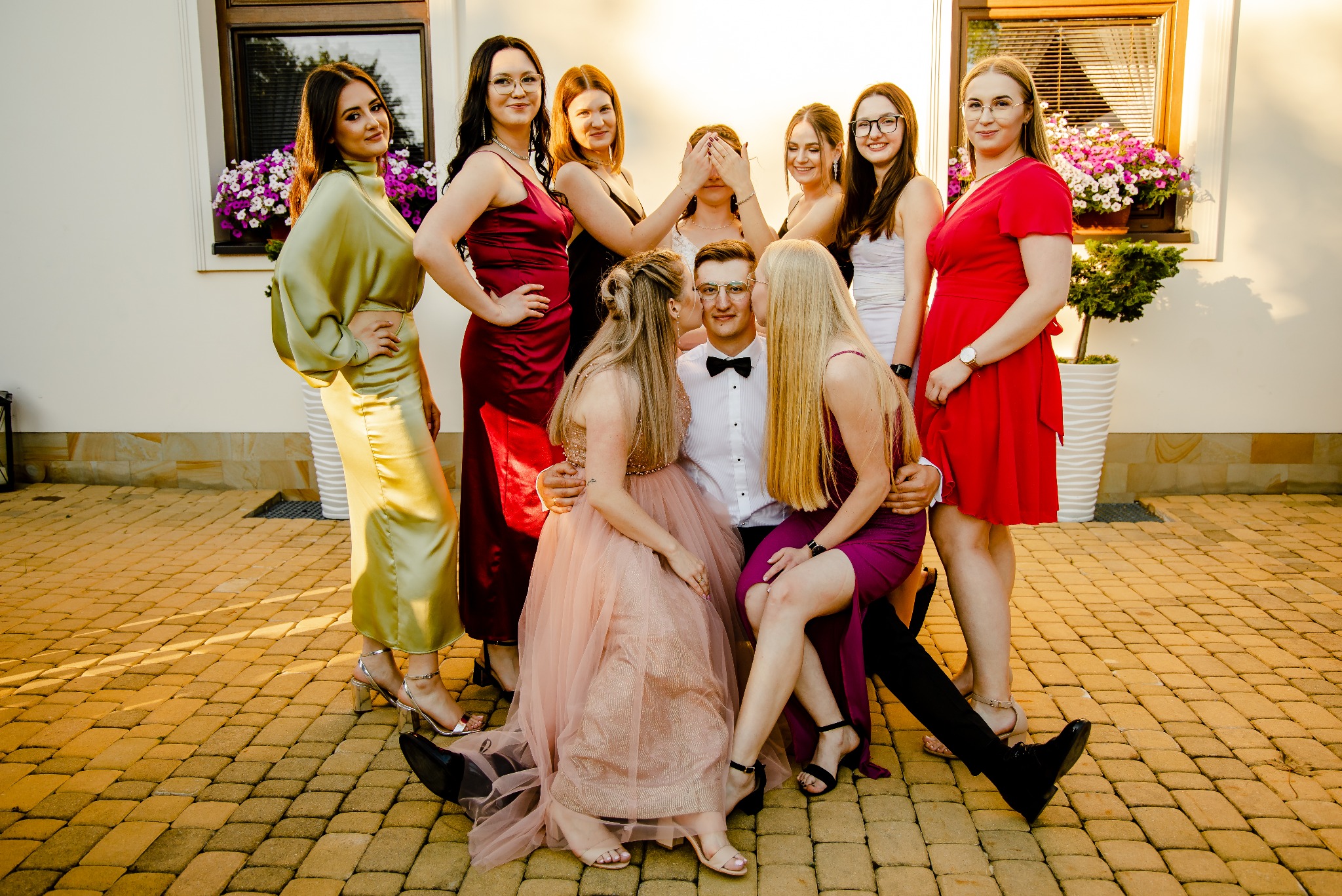 Grupa kobiet w sukienkach otacza mężczyznę w muszce, pozując na tle budynku z kwiatami w oknach, jedna z kobiet zasłania oczy mężczyzny, a dwie go całują w policzki.