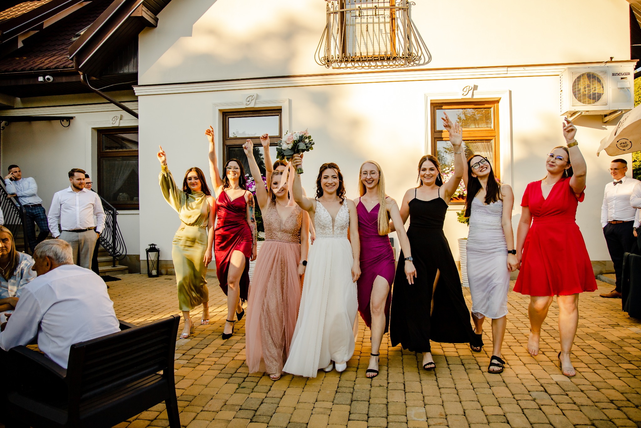 Panna młoda w białej sukni ślubnej trzyma bukiet, otoczona druhnami w kolorowych sukienkach na tle białego budynku z oknami i klimatyzacją Sinclair.