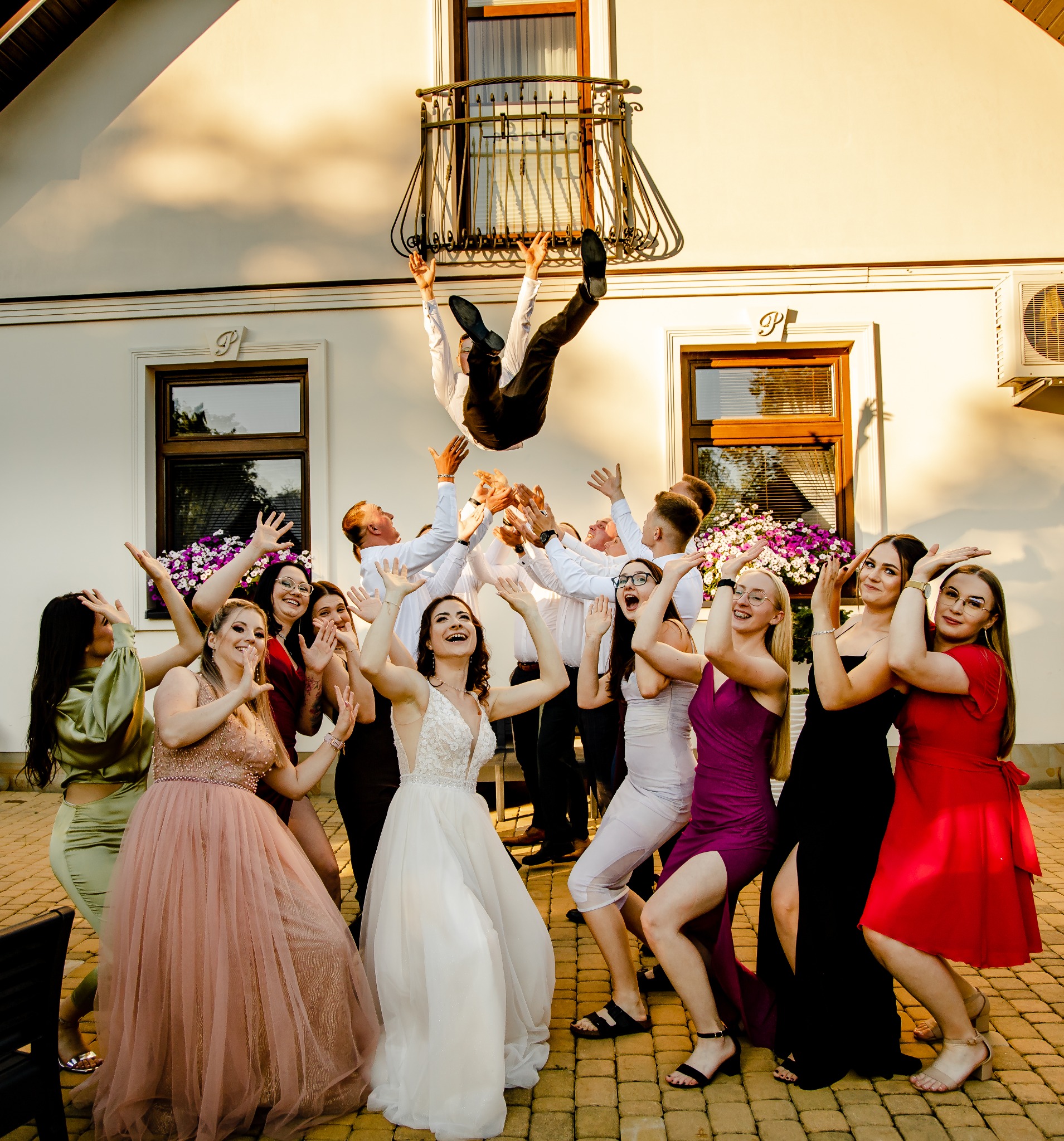 Grupa roześmianych osób w sukniach wieczorowych i koszulach podrzuca w górę mężczyznę w garniturze przed białym budynkiem z balkonem i kwiatami w oknach, radosna atmosfera.