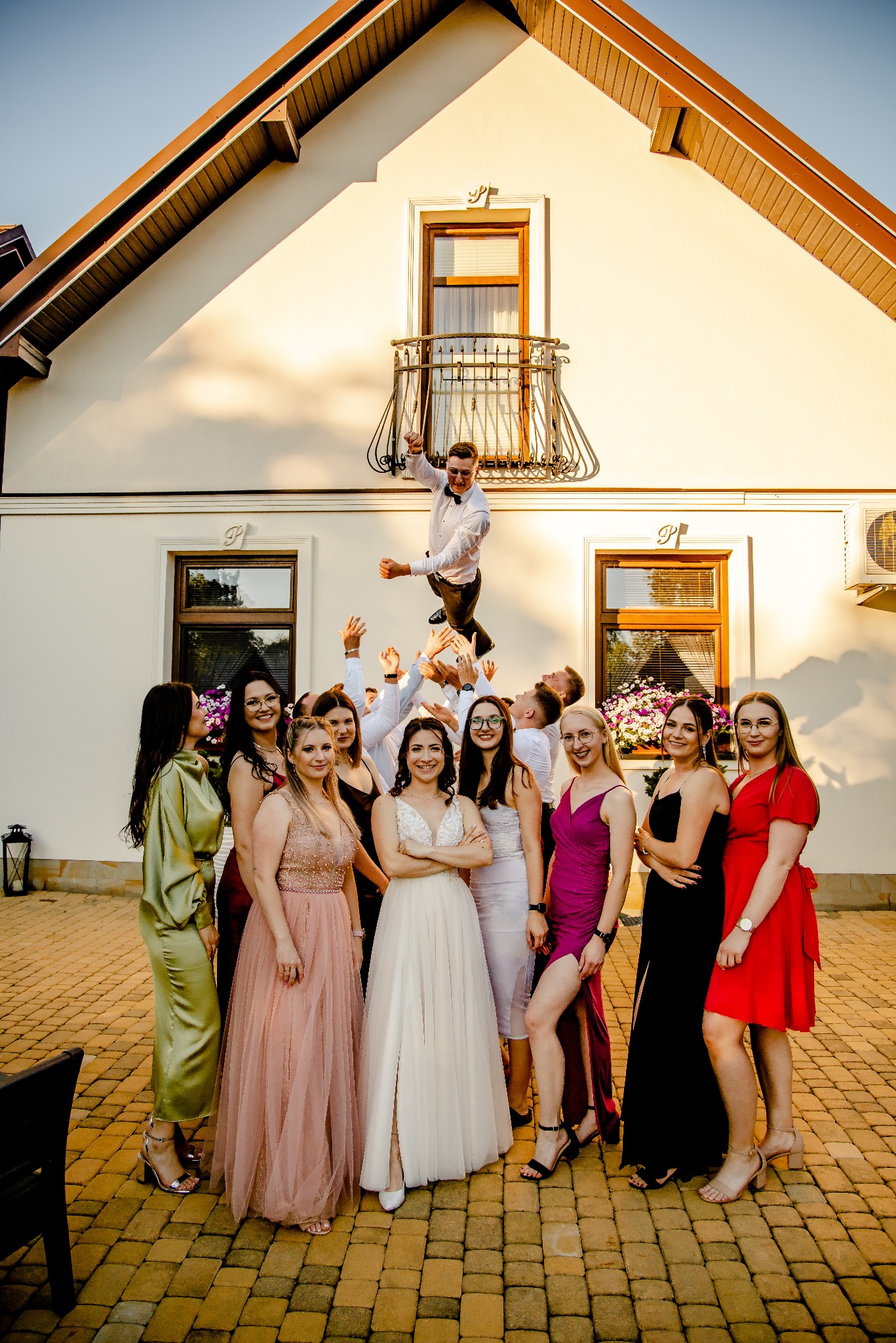 Grupa gości weselnych pozuje do zdjęcia przed budynkiem, tworząc piramidę, na której szczycie stoi mężczyzna w garniturze, z uniesionymi rękami w geście radości. Panna młoda w białej sukni stoi...