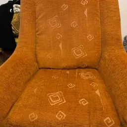 Pomarańczowy fotel z wzorem geometrycznym, widoczne ślady użytkowania i plamy na siedzisku, w tle sterta ubrań.