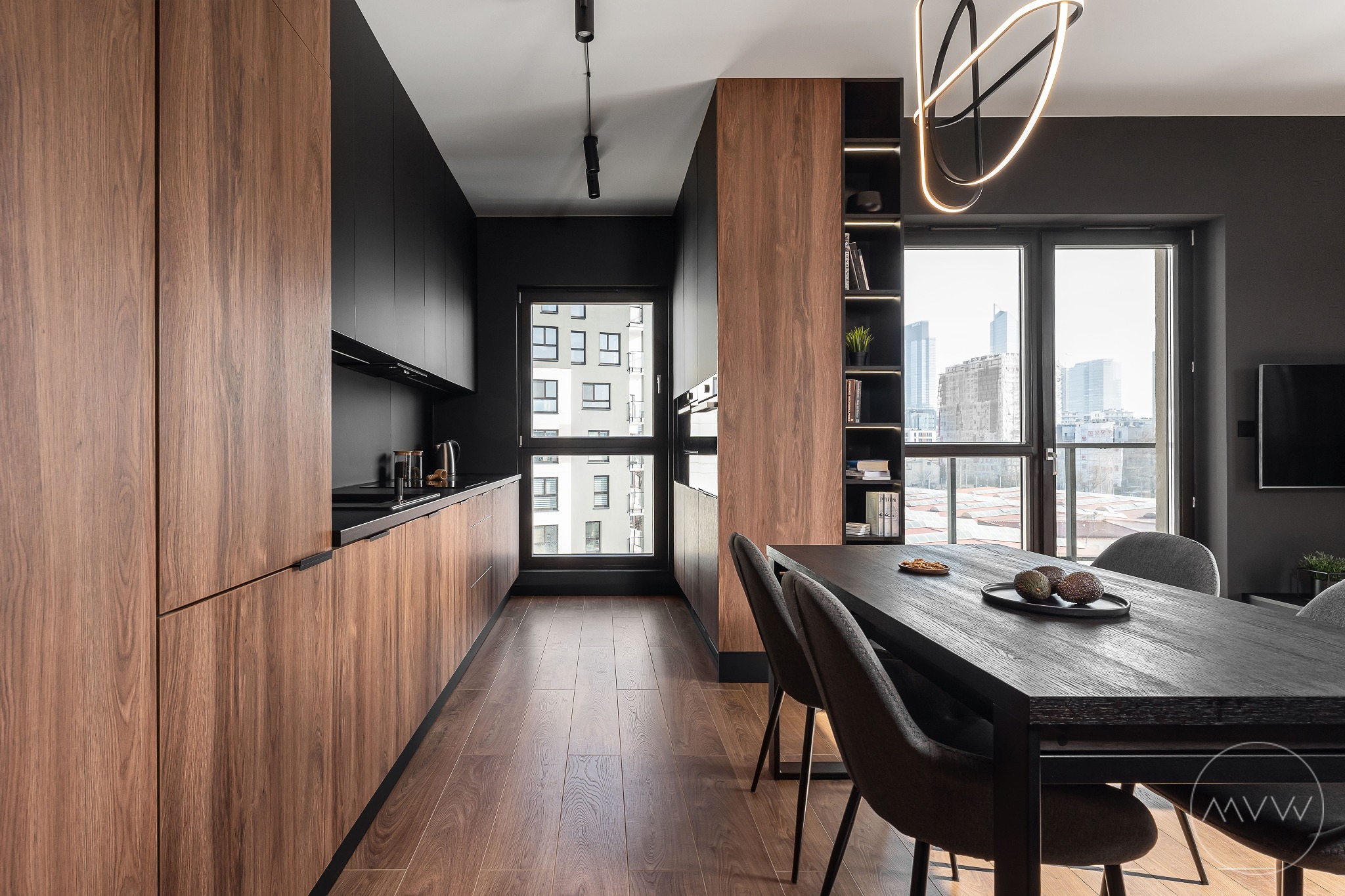 Nowoczesna kuchnia z drewnianymi szafkami dolnymi i czarnymi szafkami górnymi, uzupełniona długim stołem jadalnym z ciemnego drewna i szarymi krzesłami na tle okna z widokiem na miasto.