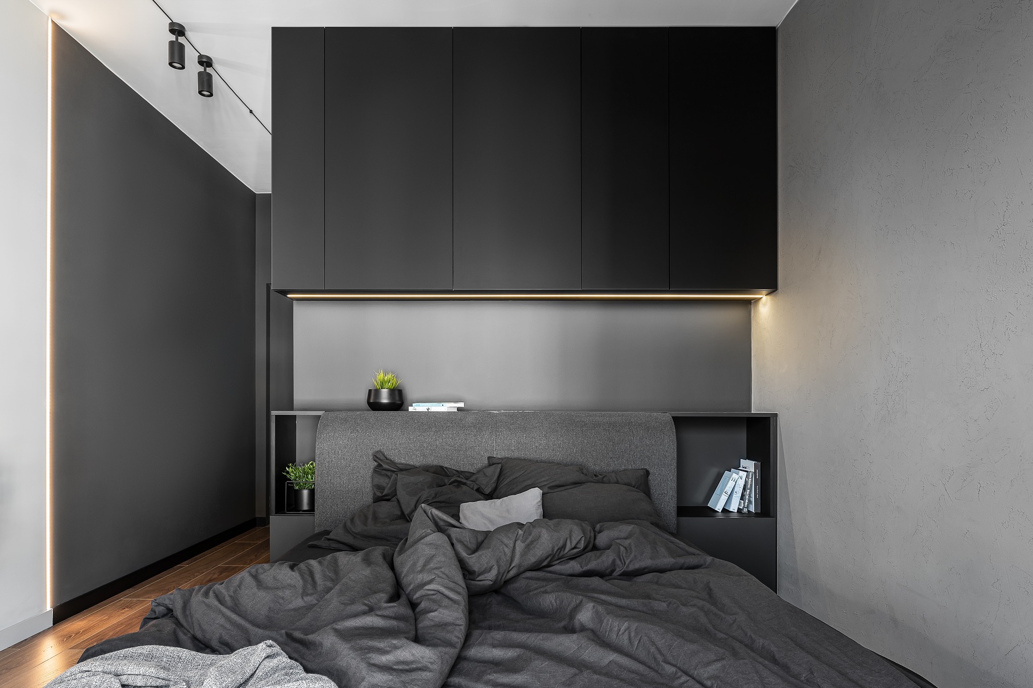 Nowoczesna sypialnia w odcieniach szarości i czerni z drewnianą podłogą, minimalistycznym oświetleniem i zabudową meblową nad łóżkiem.