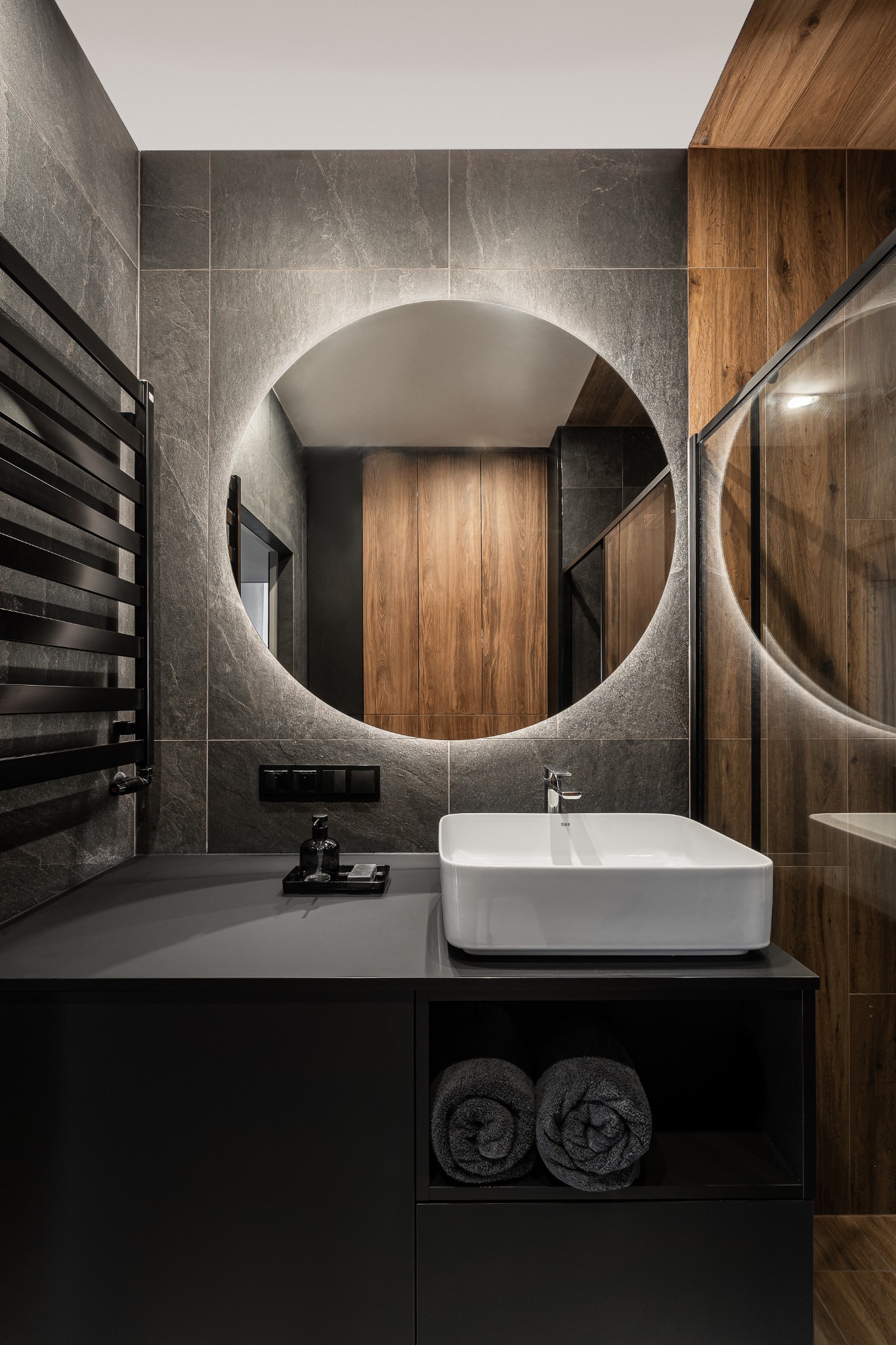 Nowoczesna łazienka z podświetlanym okrągłym lustrem, szarymi płytkami imitującymi kamień, drewnianymi panelami, czarną szafką z umywalką i ręcznikami zwiniętymi w rulony.