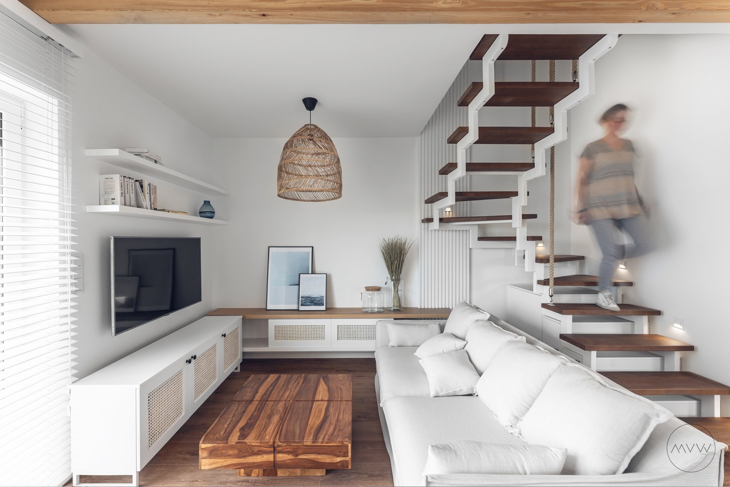 Jasne wnętrze z drewnianymi schodami ze sznurami, białą sofą, telewizorem, drewnianym stolikiem kawowym i lampą z rattanu, osoba schodząca po schodach.
