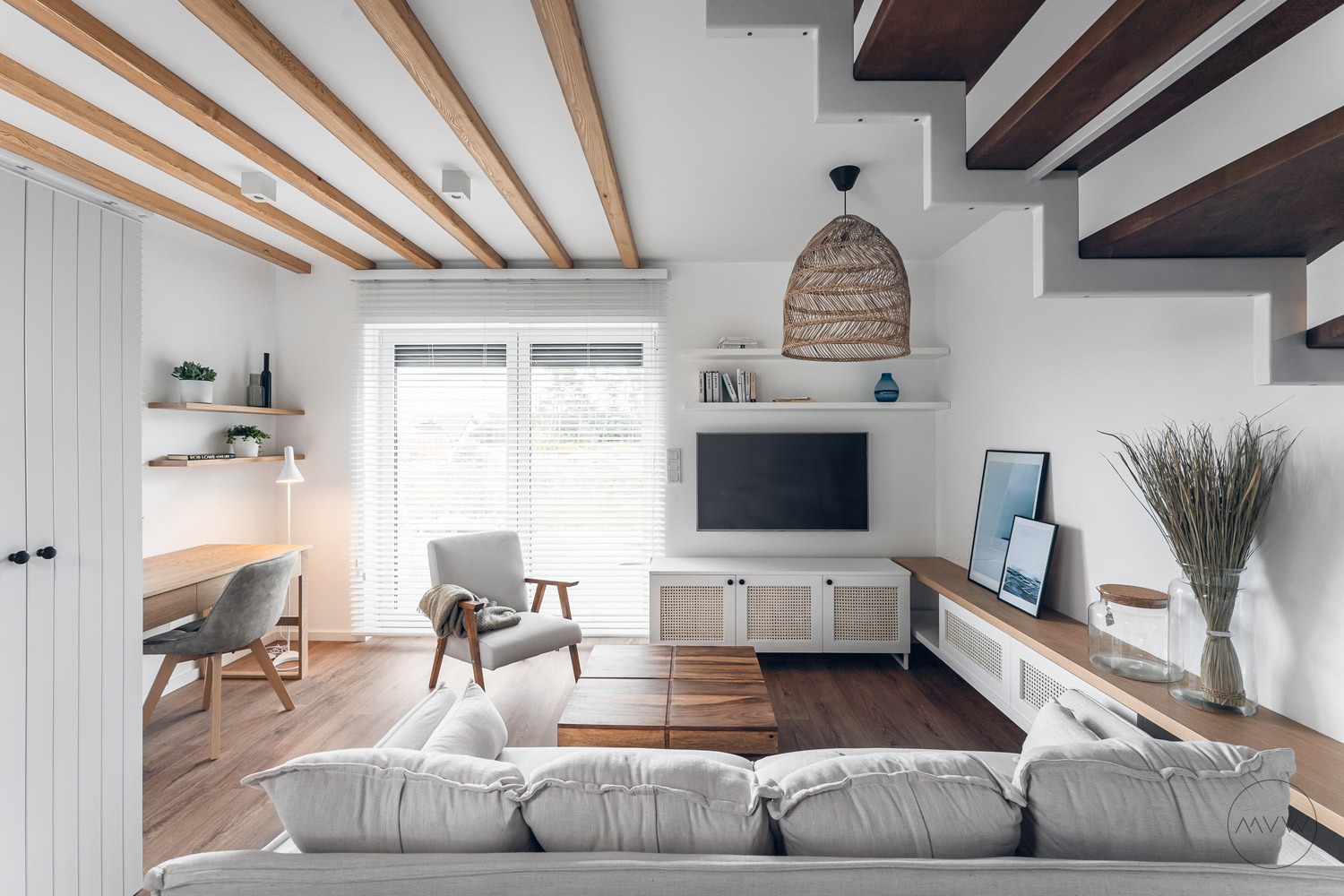 Jasne wnętrze salonu z drewnianymi belkami stropowymi, schodami z drewnianymi stopniami, szarą sofą, telewizorem na ścianie, rattanową lampą i drewnianym stolikiem kawowym.