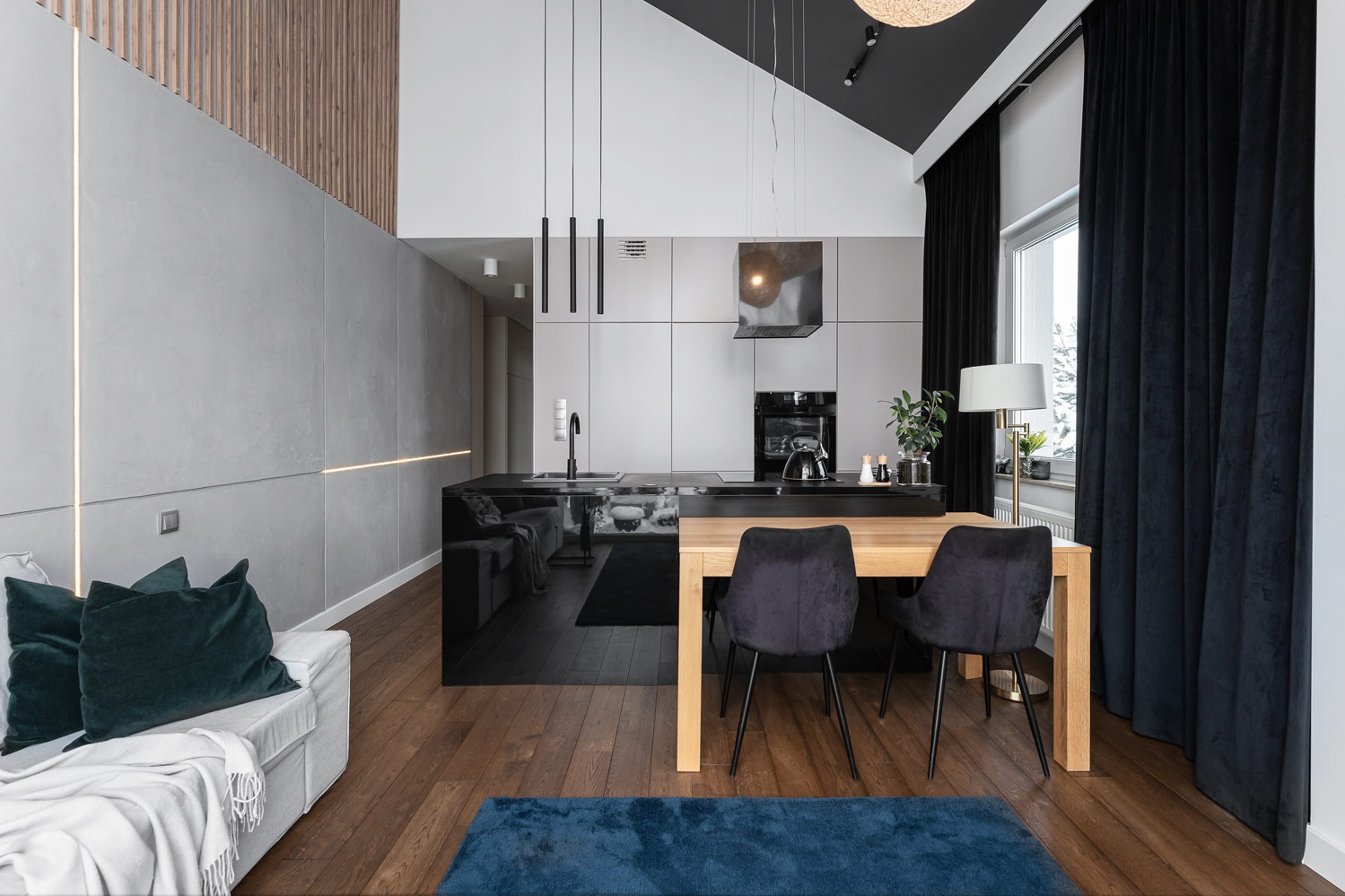 Wnętrze nowoczesnego mieszkania z otwartą kuchnią, czarnym blatem, drewnianym stołem i welurowymi krzesłami, oświetlone lampami wiszącymi i listwami LED w szarej ścianie.