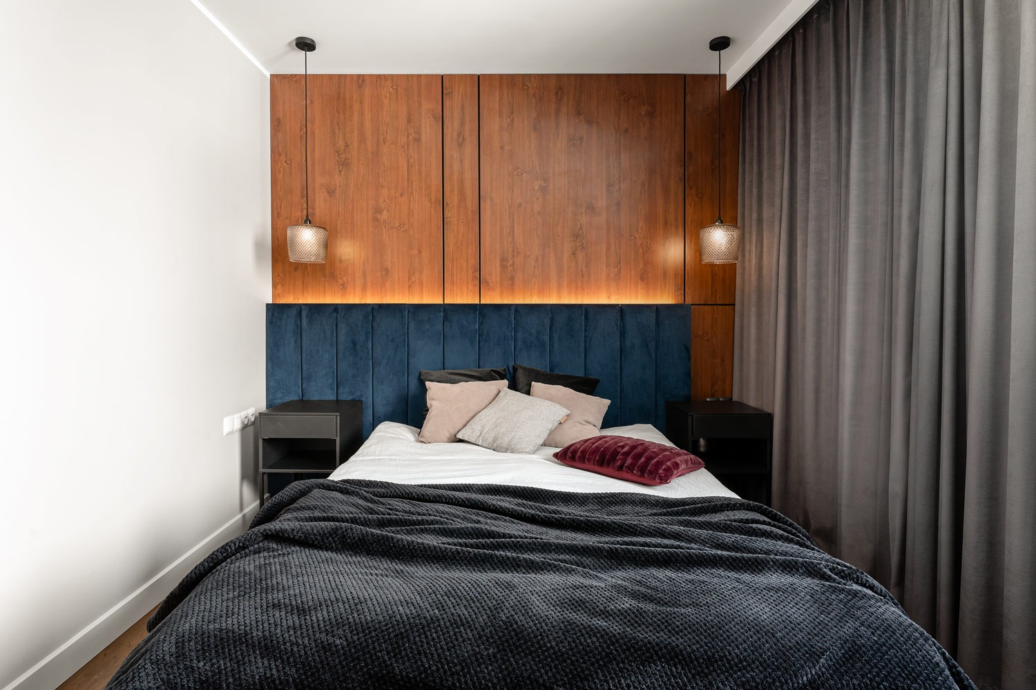 Nowoczesna sypialnia z drewnianą ścianą za łóżkiem, niebieskim welurowym zagłówkiem, szarą narzutą i lampami wiszącymi z ażurowymi kloszami.