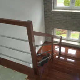 Montaż balustrady ze stali nierdzewnej i drewna na brązowych schodach, widoczne okna i ceglana ściana w tle.