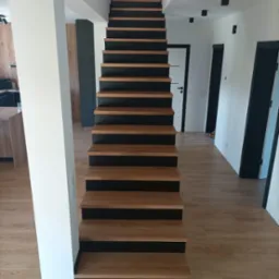 Nowoczesne schody drewniane z czarnymi podstopnicami, widok z dołu, jasne wnętrze z drewnianą podłogą i białymi ścianami, czarne drzwi w tle.