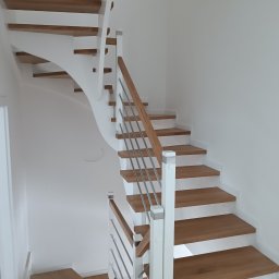 PHU DĄB-DEKOR KATARZYNA JEWULSKA - Nowoczesne schody drewniane z białymi elementami i metalową balustradą. Widok z dołu, ukazujący zakrzywiony bieg schodów i minimalistyczny design.