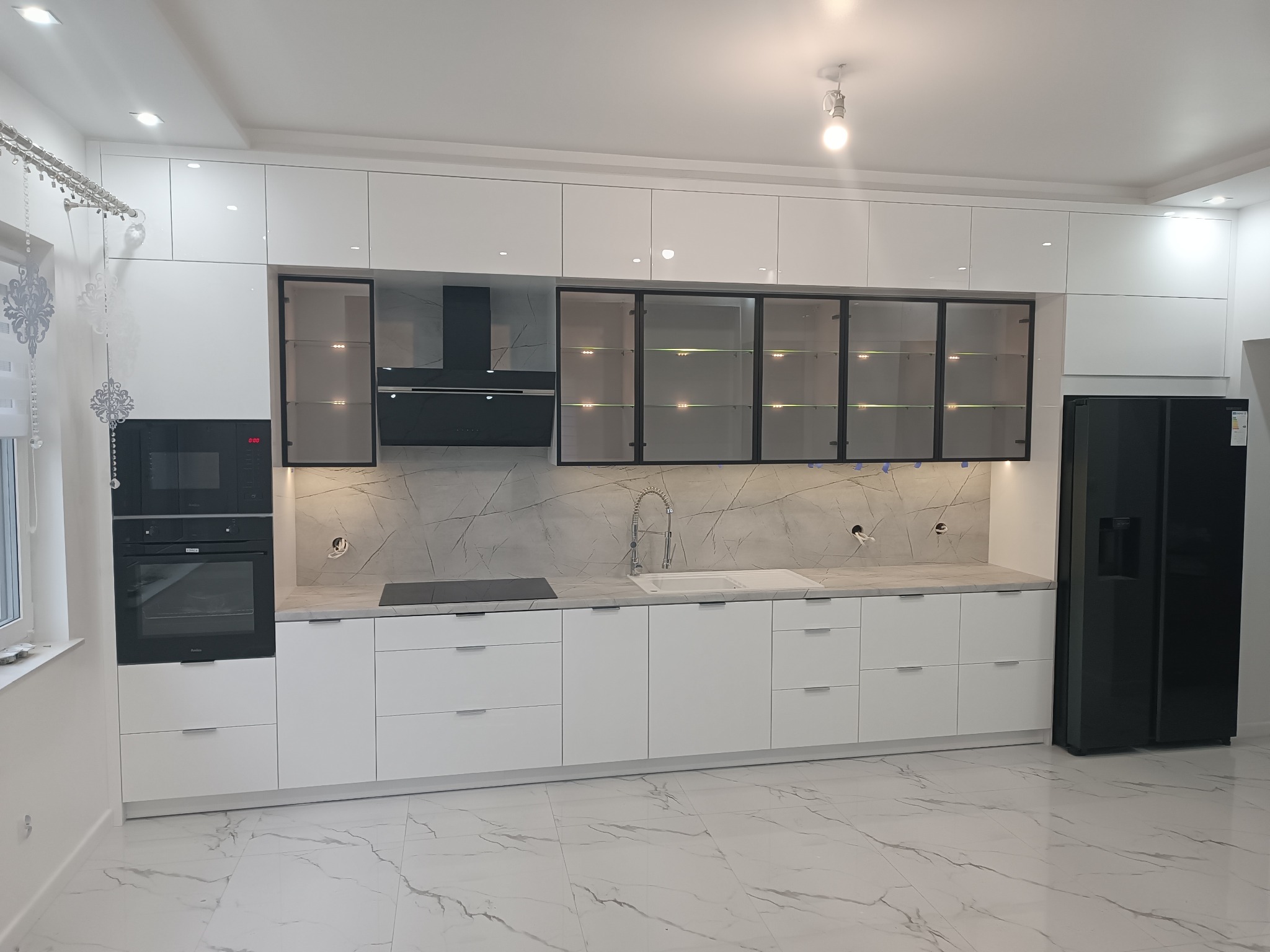 Nowoczesna, biała kuchnia z błyszczącymi frontami, szklanymi szafkami z czarną ramą i marmurkowym blatem, uzupełniona czarnym sprzętem AGD i podłogą z wzorem marmuru.