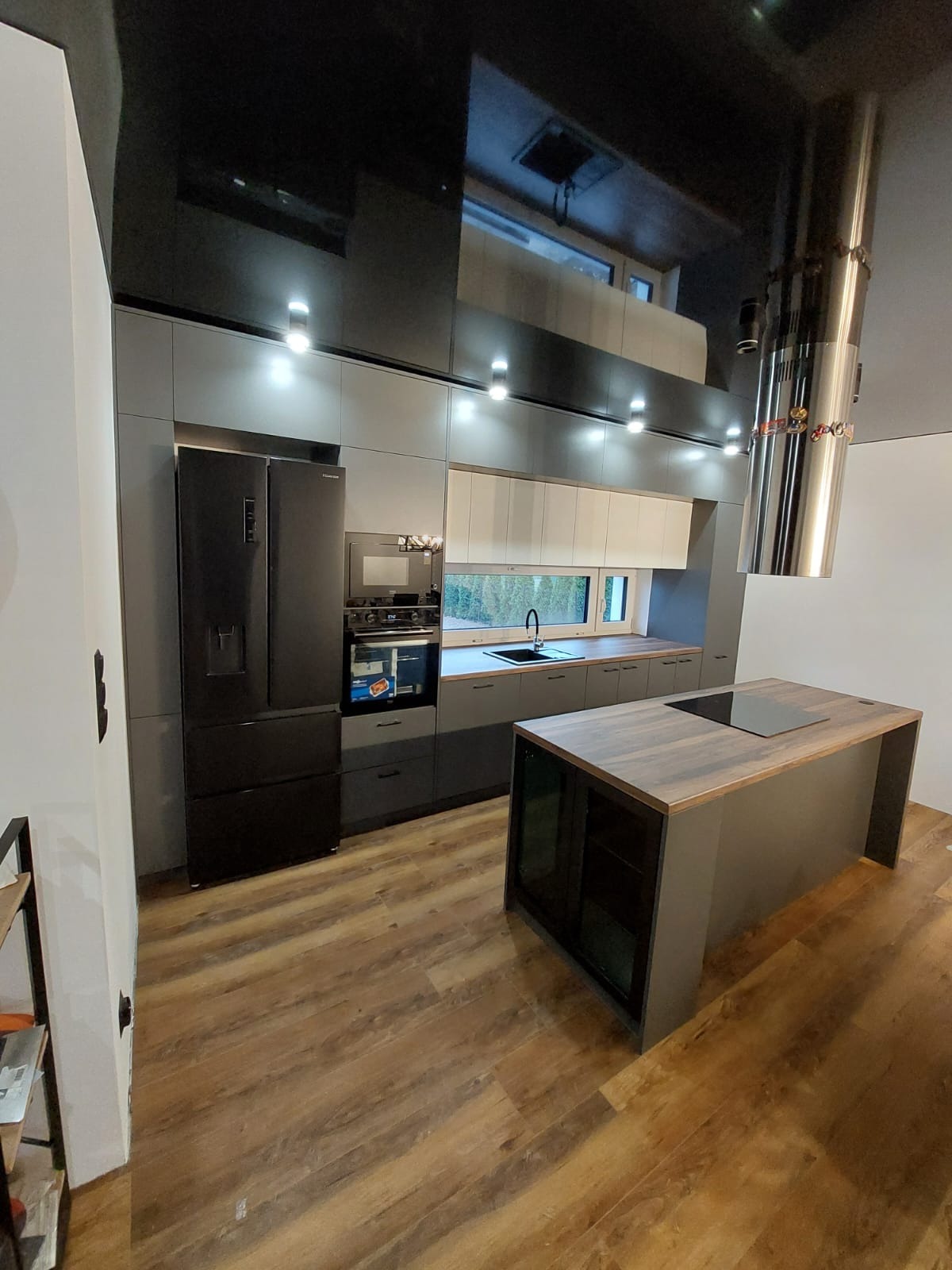 Nowoczesna kuchnia z wyspą, szarymi szafkami, czarnym sprzętem AGD i błyszczącym czarnym sufitem, oświetlona punktowymi lampami.