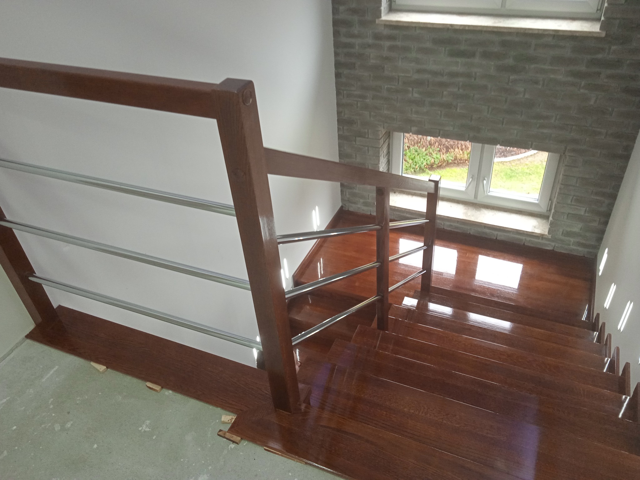Polerowane, brązowe schody drewniane z metalową balustradą, widok z góry, z elementami wykończeniowymi w trakcie montażu i oknem w tle.