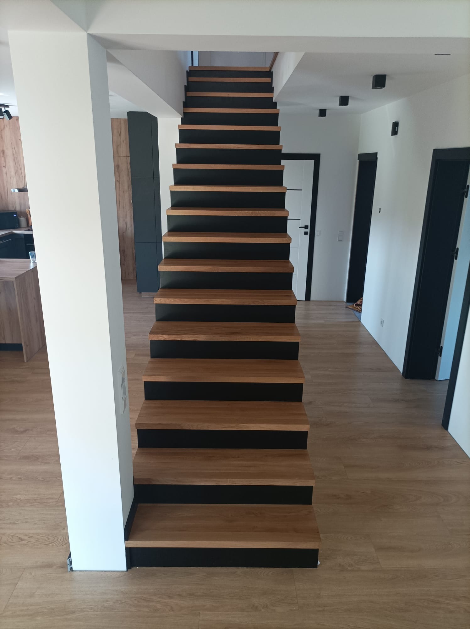 Nowoczesne schody drewniane z czarnymi podstopnicami, widok z dołu, jasne wnętrze z drewnianą podłogą i białymi ścianami, czarne drzwi w tle.