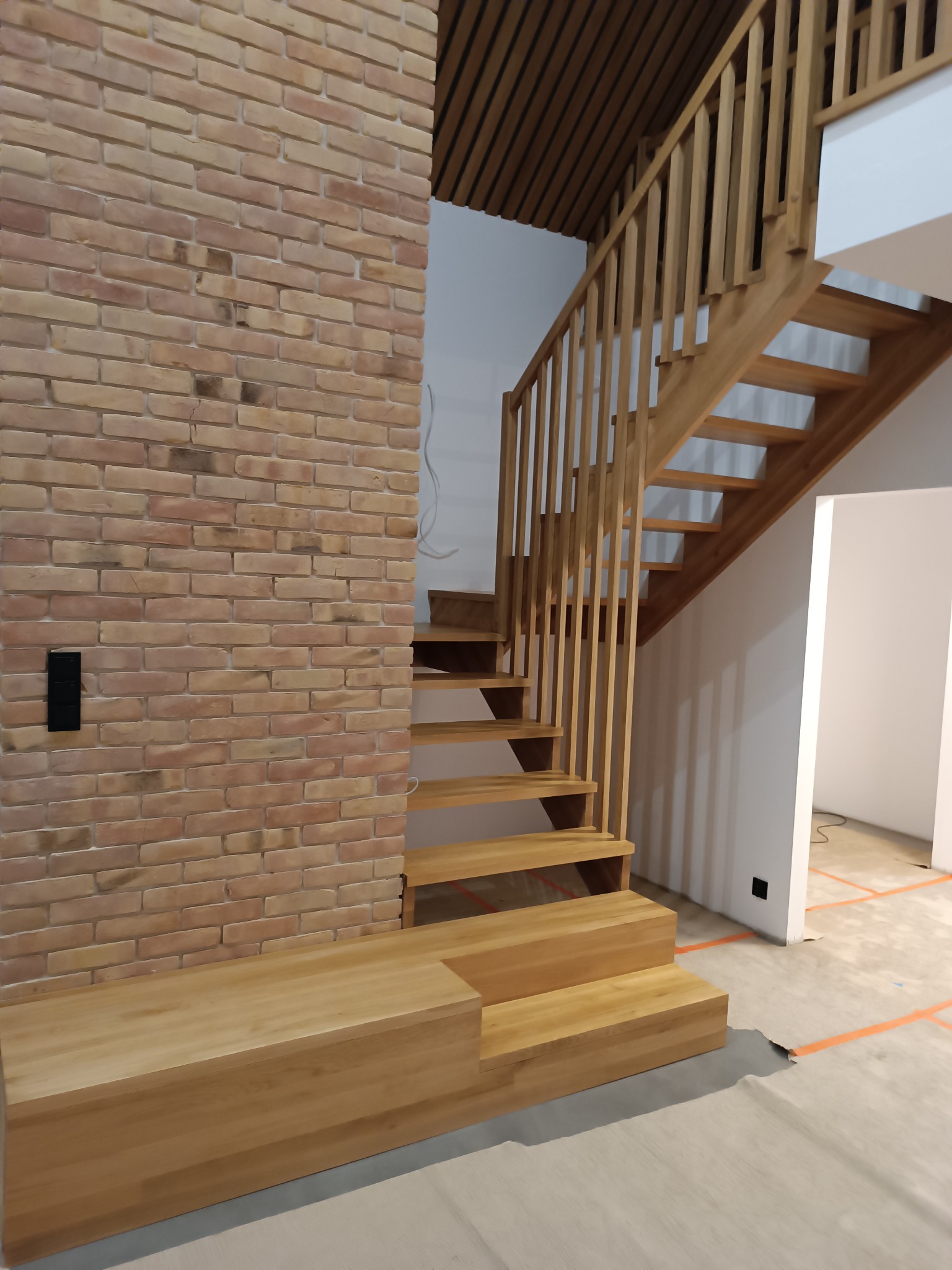 Jasne schody drewniane z balustradą, ceglana ściana z czarnym włącznikiem, drewniany sufit. Widok na białe pomieszczenie z pomarańczowymi liniami na podłodze.