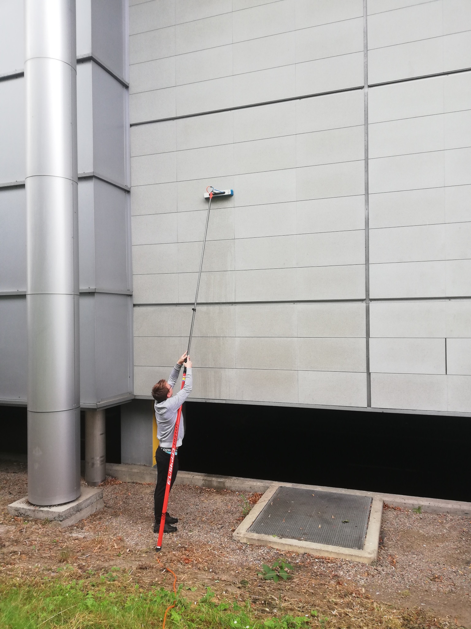 Pracownik myje elewację budynku przemysłowego za pomocą teleskopowej lancy z szczotką, widoczne ślady spływającej wody na jasnej elewacji.