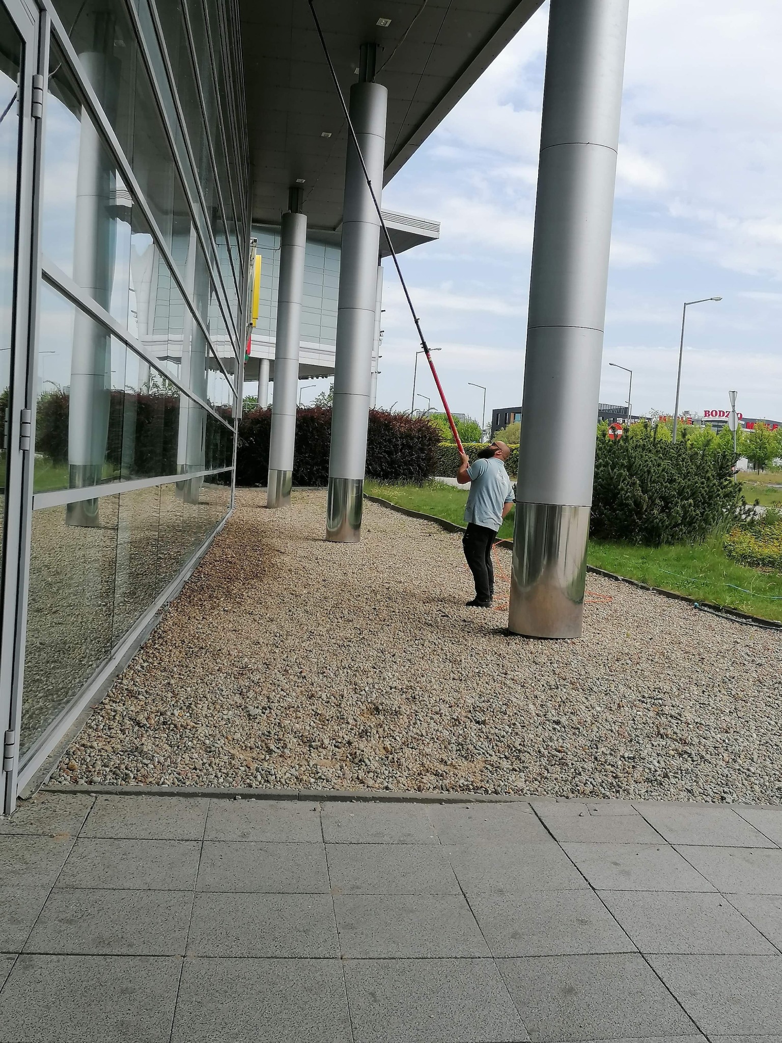 Pracownik myje szklaną elewację budynku biurowego za pomocą teleskopowego kija z zamocowaną szczotką, widoczne odbicia w szybach i otoczenie budynku.