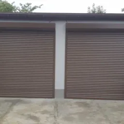 Dwie zamknięte, brązowe bramy garażowe segmentowe z poziomymi panelami, widoczne z zewnątrz, z workami kory i cegłami obok prawej bramy.